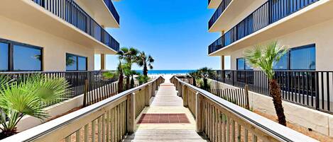 A wooden pathway leading to a beach with a clear sky, surrounded by buildings with balconies.