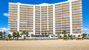Een hotel met meerdere verdiepingen en balkons, gelegen aan een strand met palmbomen.