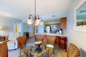 A dining area with a glass table, wicker chairs, a chandelier, and a kitchen in the background. - Serene water view condo with pools, sauna, tennis court, fitness center, W&D (Orange Beach)