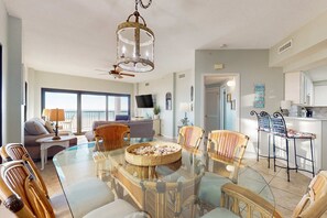 A spacious living area with a glass dining table, wicker chairs, a ceiling fan, and a large window. - Delightful Corner Getaway With Gulf Views, Beach Access, Free WiFi & Shared Pools (Orange Beach)