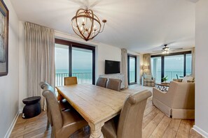 A spacious living room with a large wooden dining table, comfortable seating, a chandelier, a television, and large windows offering a view of the sea. - Lovely resort condo with spectacular Gulf views, pool & tennis (Orange Beach)