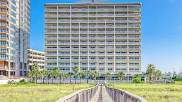 A multi-story hotel with a central atrium, a wooden boardwalk leading to the entrance, and palm trees in the foreground.