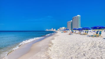 A beach with white sand, a clear blue sky, and a tall hotel building in the background.