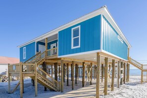 Un bâtiment en bois bleu, doté d’un escalier et d’une terrasse en bois, surélevé sur des piliers, situé sur une plage de sable.