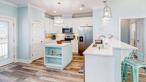 Uma cozinha moderna com armários brancos, uma ilha central, eletrodomésticos de aço inoxidável e cadeiras cor de azul-turquesa.