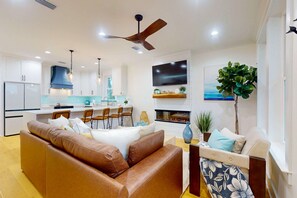 Ein modernes Wohnzimmer mit einem braunen Ledersofa, einem weißen Sofa, einem Essbereich mit Stühlen, einer Küche mit weißen Schränken, einem Deckenventilator, einem Flachbildfernseher, einem Kamin und einer großen Pflanze.
