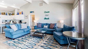 Una sala de estar con un sofá azul, una mesa de centro y una pequeña mesa redonda. Hay una zona de cocina con armarios blancos y una mesa de comedor. La habitación tiene techos altos con vigas a la vista y un gran ventanal con cortinas.