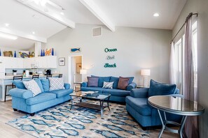 Ein Wohnzimmer mit blauem Sofagarnitur, Couchtisch und kleinem runden Tisch. Es gibt eine Küchenzeile mit weißen Schränken und einem Esstisch. Der Raum hat eine hohe Decke mit freiliegenden Balken und ein großes Fenster mit Vorhängen.