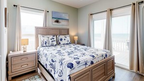 Un dormitorio con cama, mesitas de noche, ventilador de techo, ventana con cortinas y una decoración mural con temática de playa.