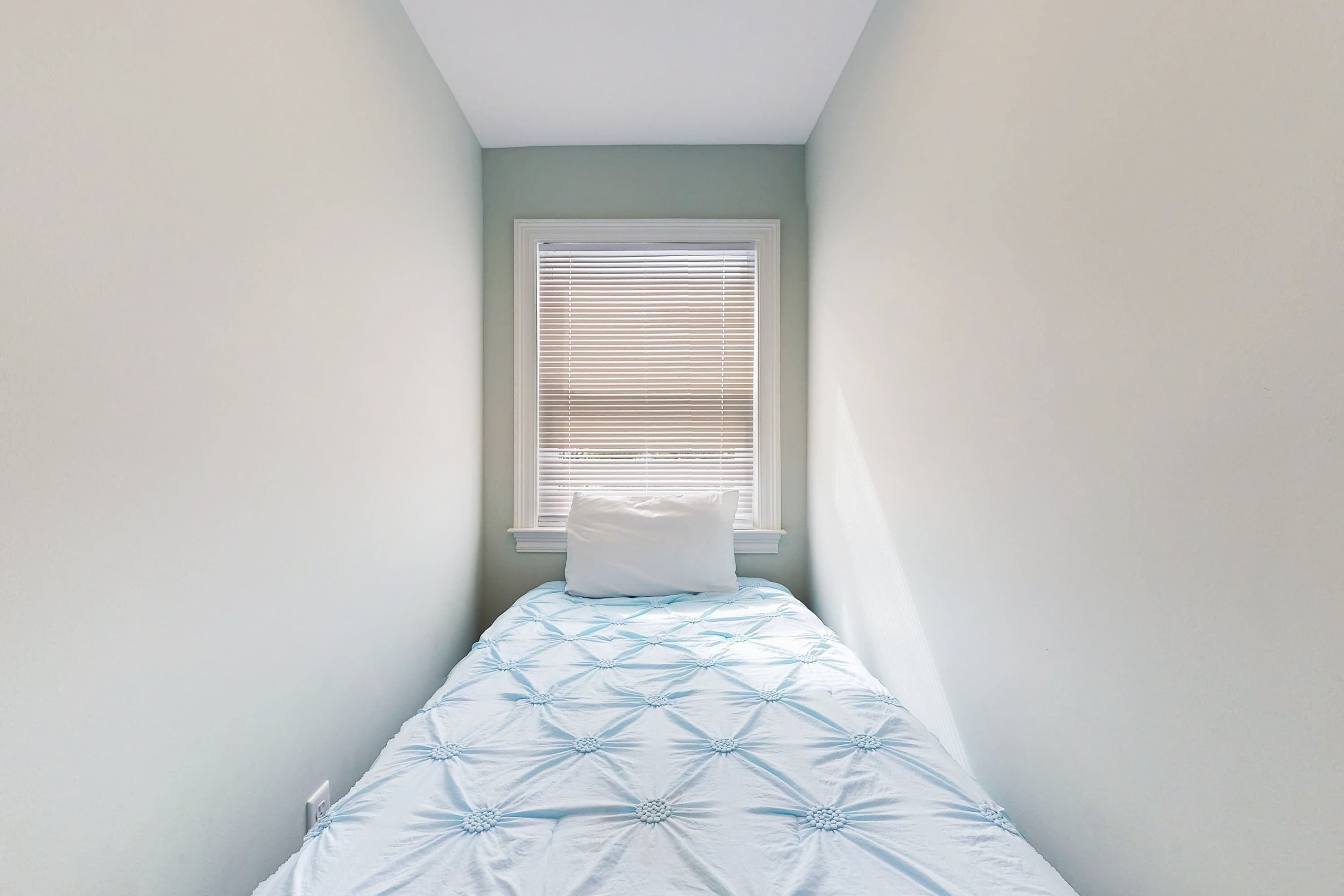 Ein schmales Zimmer mit einem Einzelbett, einem Fenster mit Jalousien und einer hellgrünen Wand.