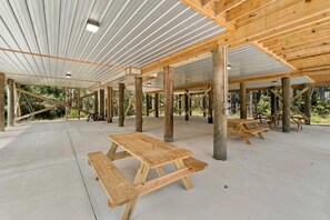 Un pavillon ouvert, doté de bancs en bois et d’un sol en béton, entouré d’arbres.