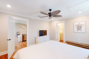 Une chambre à coucher avec un lit, un ventilateur de plafond, un téléviseur posé sur une commode, une porte de salle de bain et un tableau encadré accroché au mur.