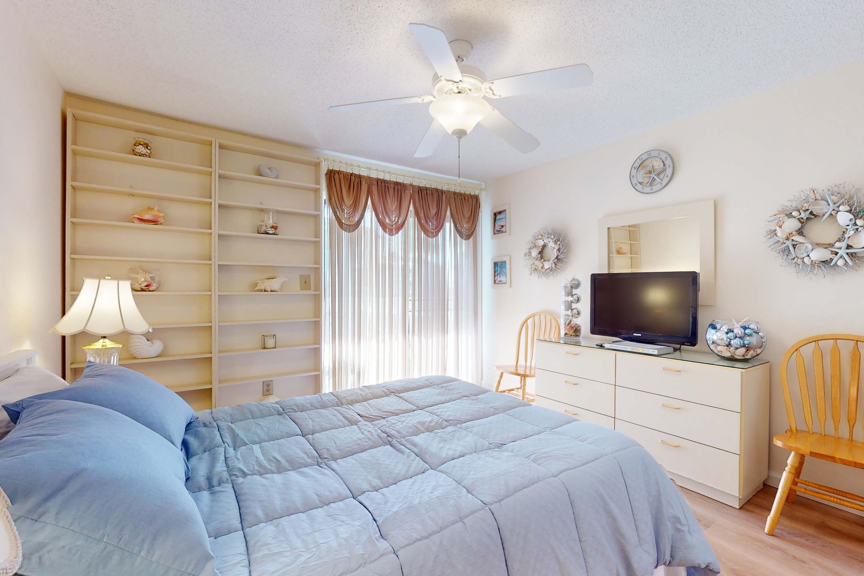 A bedroom with a bed, a TV, a wardrobe, a ceiling fan, and a window with curtains.