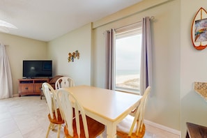 A room with a dining table, chairs, a TV, and a view of the beach. - Cozy Condo in Gulf-Front Complex w/Free WiFi, Shared Pool, Central AC, Elevator (Gulf Shores)