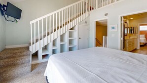 Quarto com cama, escada com corrimão branco, televisão na parede e banheiro visível através de uma porta aberta.