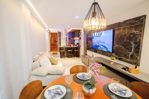 A modern dining area with a wooden table set for two, a TV mounted on a marble wall, and a cozy seating area in the background. - Atlantis #302 - Ape em Cabo Branco por Carpediem (João Pessoa)