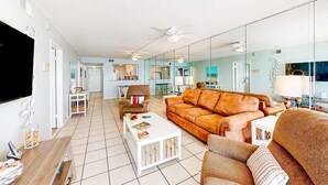 A living room with a sofa, coffee table, and a TV. There is a kitchen in the background. - 8th-floor beachfront condo with Gulf view, waterfront pool, & balcony (Gulf Shores)