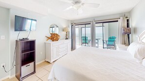 A hotel room with a bed, a ceiling fan, a TV, a dresser, a mirror, and a balcony with chairs. - 8th-floor beachfront condo with Gulf view, waterfront pool, & balcony (Gulf Shores)