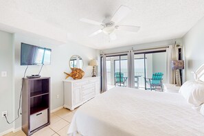 A hotel room with a bed, a ceiling fan, a TV, a dresser, a mirror, and a balcony with chairs. - 8th-floor beachfront condo with Gulf view, waterfront pool, & balcony (Gulf Shores)