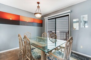 A dining area with a glass table and wicker chairs, a ceiling light, and a sliding glass door. - Comfortable Condo at a Beachfront Complex w AC, WiFi, Balconies & Shared Pool (Gulf Shores)