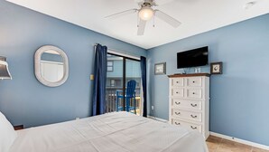 Une chambre à coucher comprenant un lit, un ventilateur de plafond, une télévision, une commode et une fenêtre avec des rideaux.