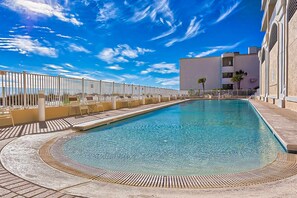 A swimming pool with a curved edge, surrounded by a white fence, and a building with balconies in the background. - Lovely Gulf-front condo with pool, sauna, washer dryer, & central AC (Gulf Shores)