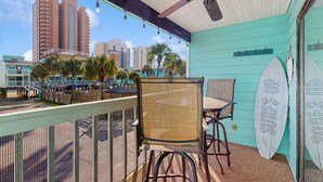 A balcony with chairs, a surfboard, and a view of tall buildings. - Beachview condo with community pools, grills, and internet (Gulf Shores)