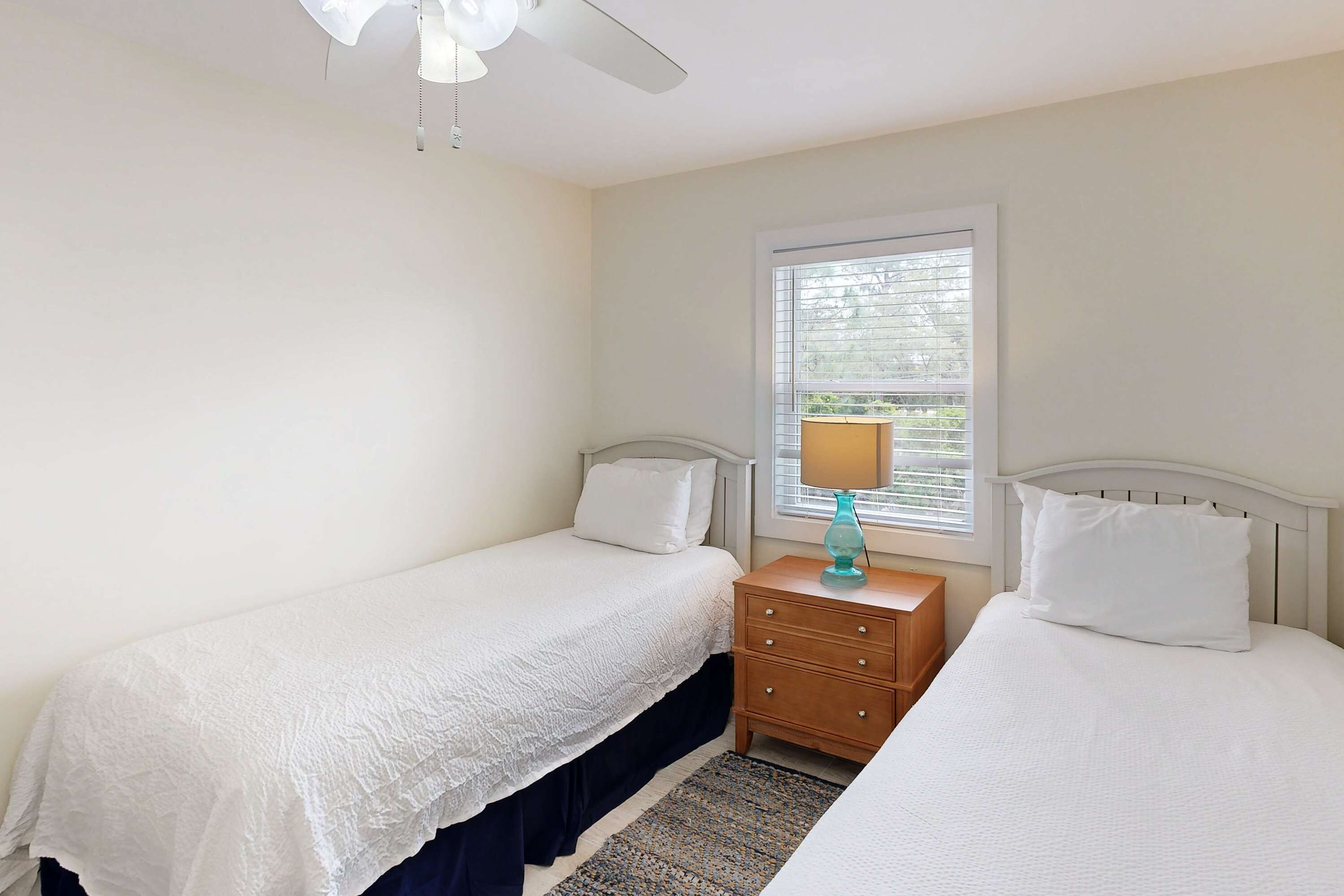 A bedroom with two beds, a nightstand, a lamp, a ceiling fan, and a window with blinds.