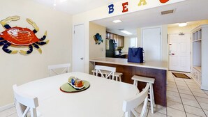 A dining area with a white table and chairs, a crab wall art, and a kitchen in the background. - Sunny gulf-front condo with private balcony, shared outdoor pool, great views (Gulf Shores)