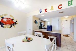 Un comedor con una mesa y sillas blancas, una obra de arte de cangrejo en la pared y una cocina al fondo.