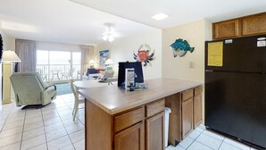 A kitchen with a refrigerator, a dining table, a chair, and a window. - Sunny gulf-front condo with private balcony, shared outdoor pool, great views (Gulf Shores)