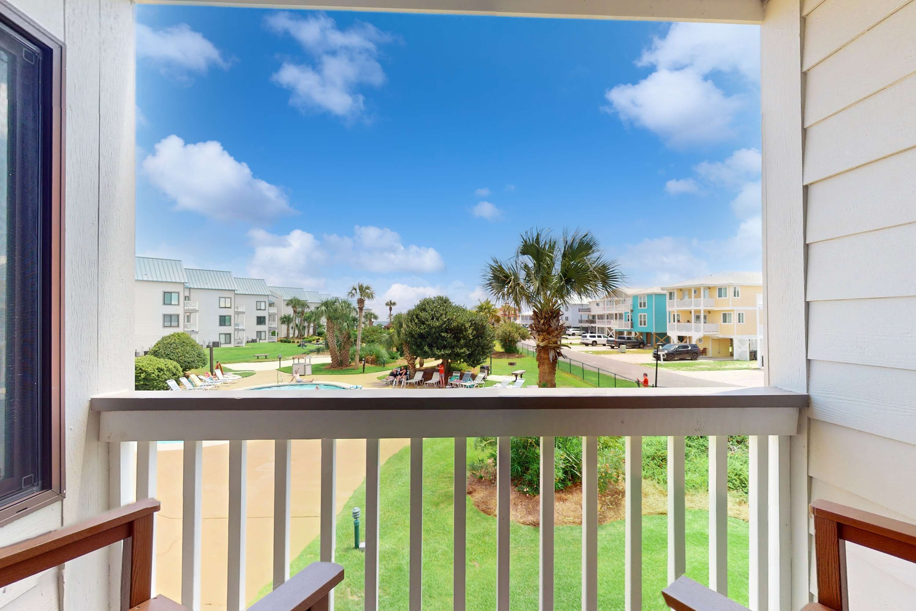 A balcony with a view of a pool area, palm trees, and multi-story buildings in the background.