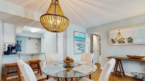 A dining area with a glass table, white upholstered chairs, a wooden floor, and a chandelier.
