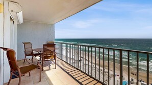 A balcony with a table and chairs, overlooking a beach with waves and people.