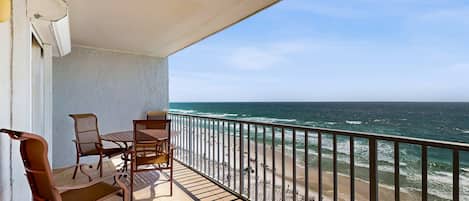 Un balcone con tavolo e sedie, vista su una spiaggia con onde e persone.