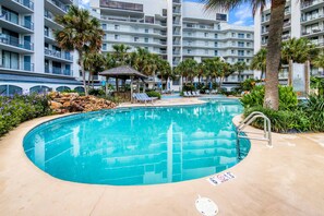 A swimming pool with lounge chairs, palm trees, and a multi-story building with balconies. - Lagoon front condo with outdoor pool, pickleball court, and piers (Gulf Shores)
