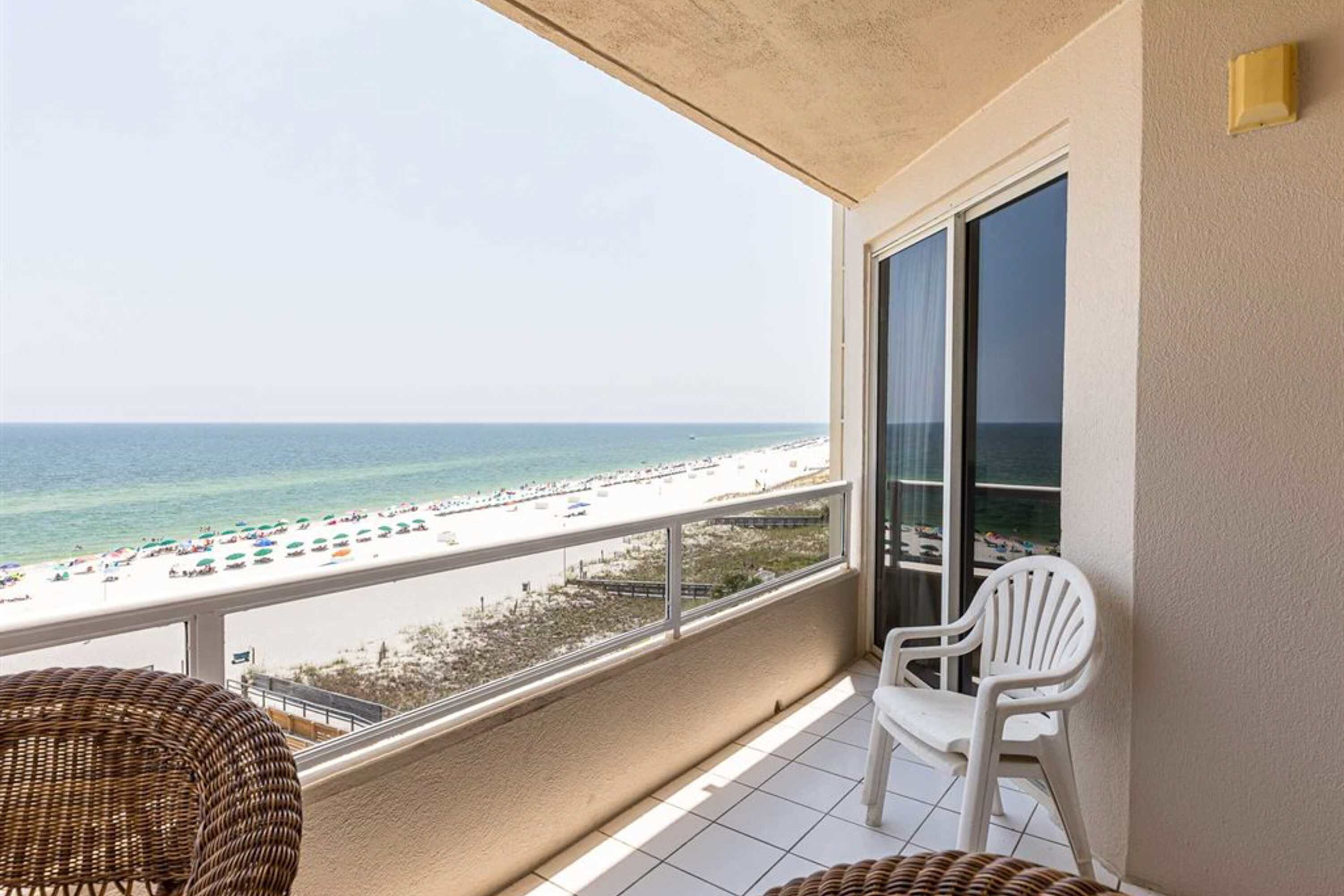 Ein Balkon mit Blick auf den Strand, ein Korbstuhl, ein weißer Plastikstuhl und ein großes Fenster.