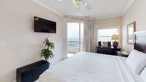 Una camera d'albergo con un letto, una ventola a soffitto, una televisione, una pianta, una finestra con tende, un comodino e una lampada.