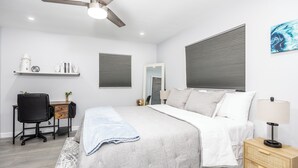 Een slaapkamer met een bed, een bureau, een stoel, een nachtkastje, een lamp, een plafondventilator en een raam met jaloezieën.