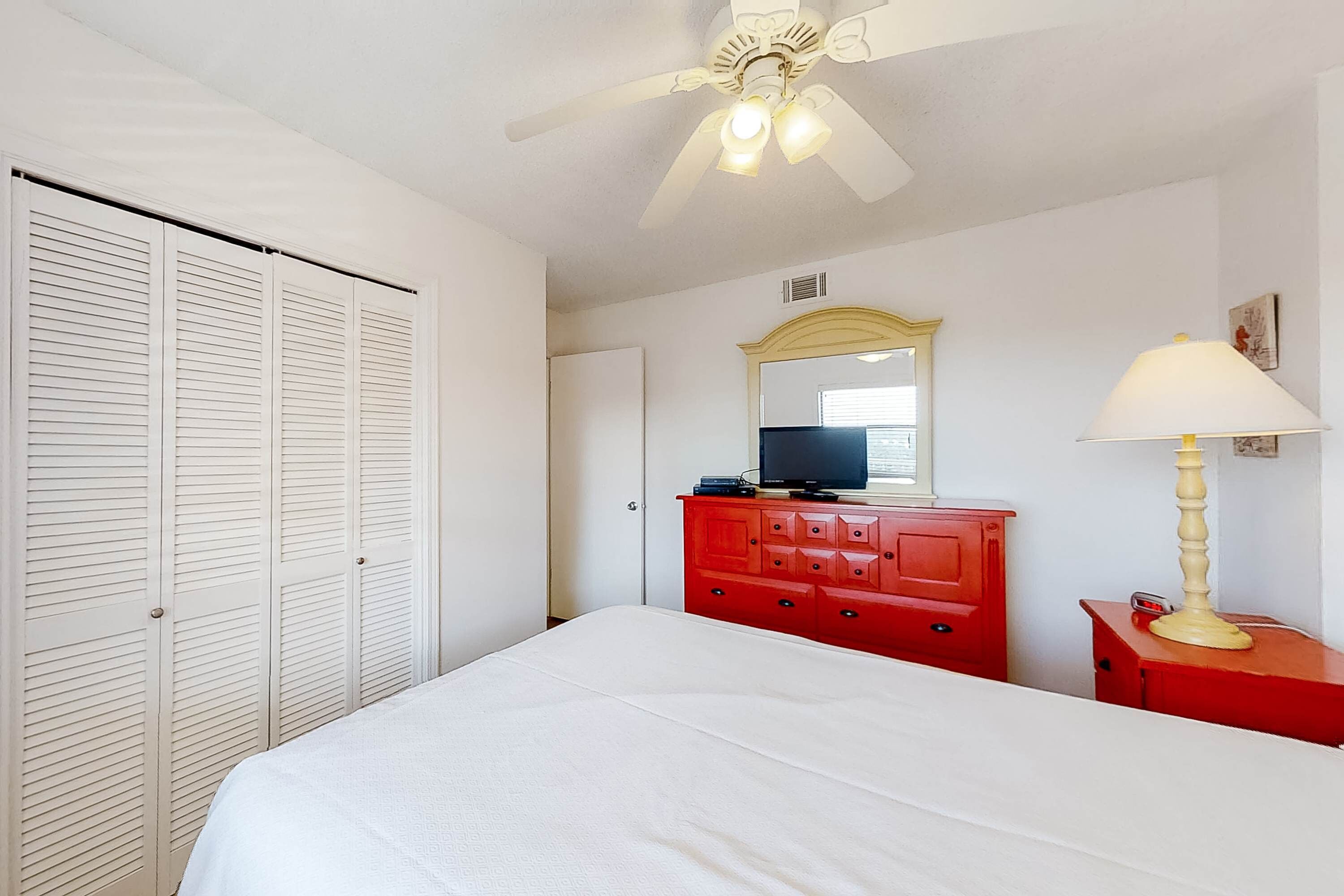 A bedroom with a bed, a red dresser, a television, and a ceiling fan.