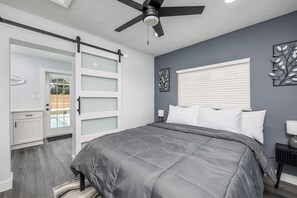 Une chambre à coucher comprenant un lit, un ventilateur de plafond, une fenêtre avec des stores, une porte avec des panneaux en verre dépoli et une petite table de chevet avec une lampe.