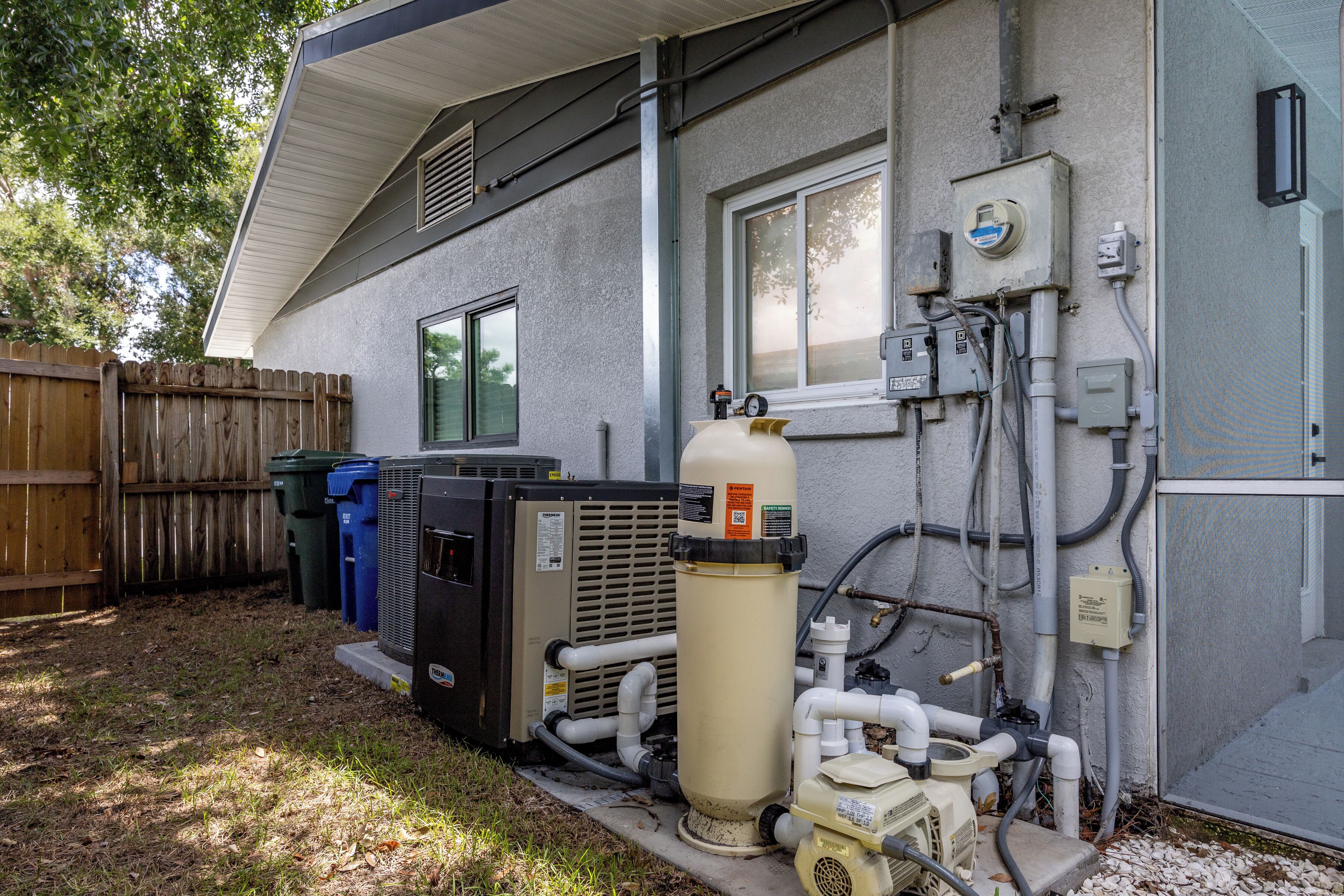 An outdoor utility area with HVAC units, a water pump, and various pipes and electrical boxes.