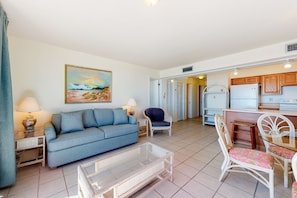 A living room with a sofa, coffee table, and chairs, a kitchen area with cabinets and appliances, and a painting on the wall. - Fantastic beachside condo with pool, sauna, WiFi & central AC (Orange Beach)