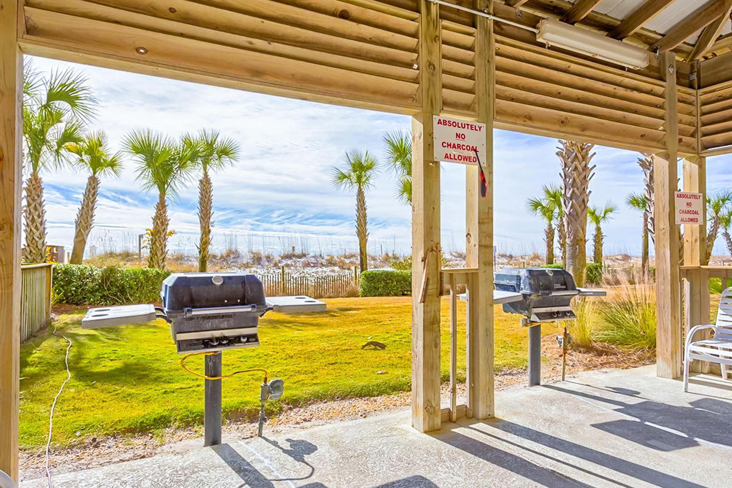 A covered outdoor area with barbecue grills, palm trees, and a sign stating 'ABSOLUTELY NO CHARCOAL ALLOWED.'