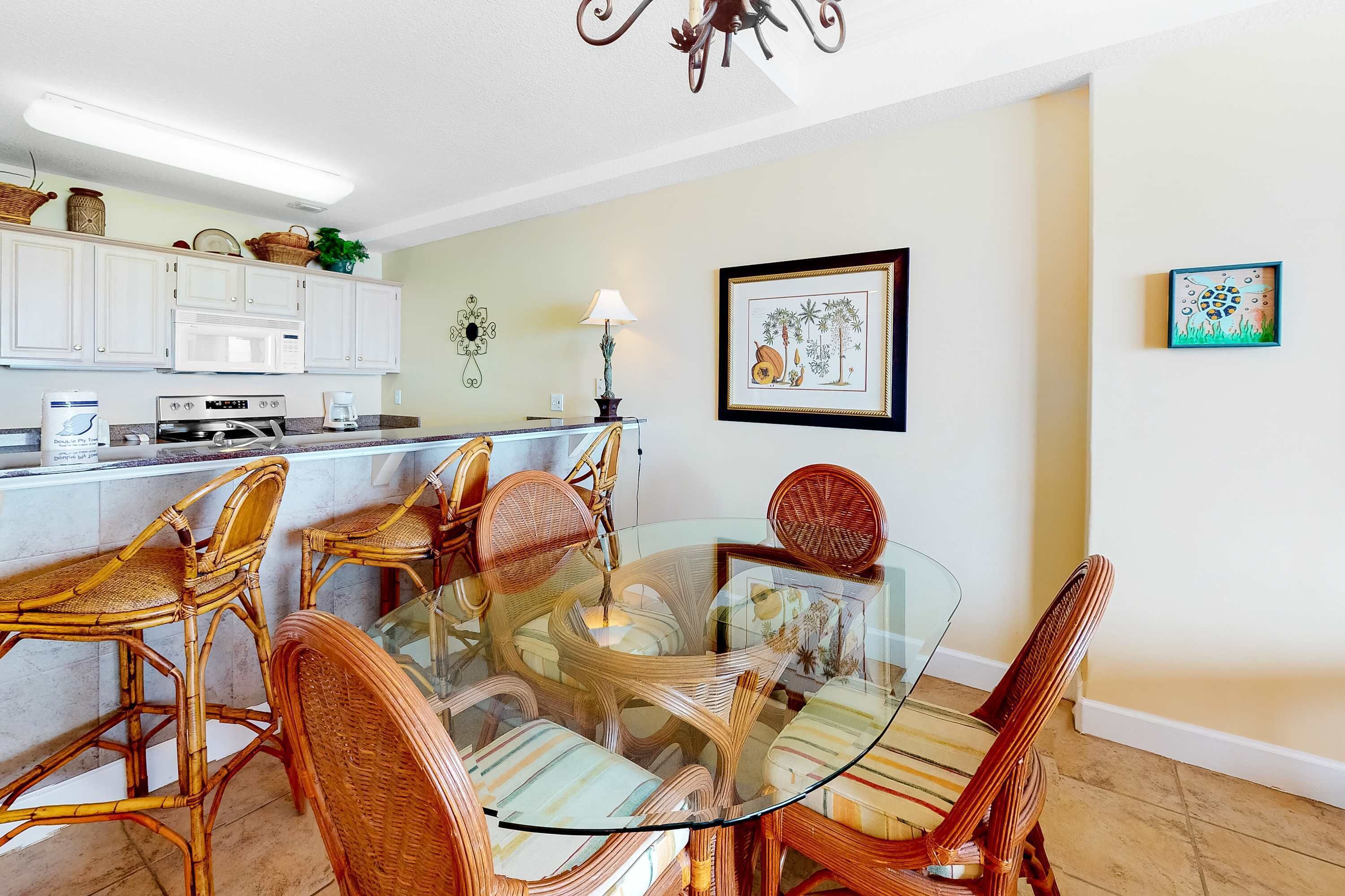 A dining area with wicker chairs and a glass table, kitchen cabinets, a microwave, and a framed picture on the wall.