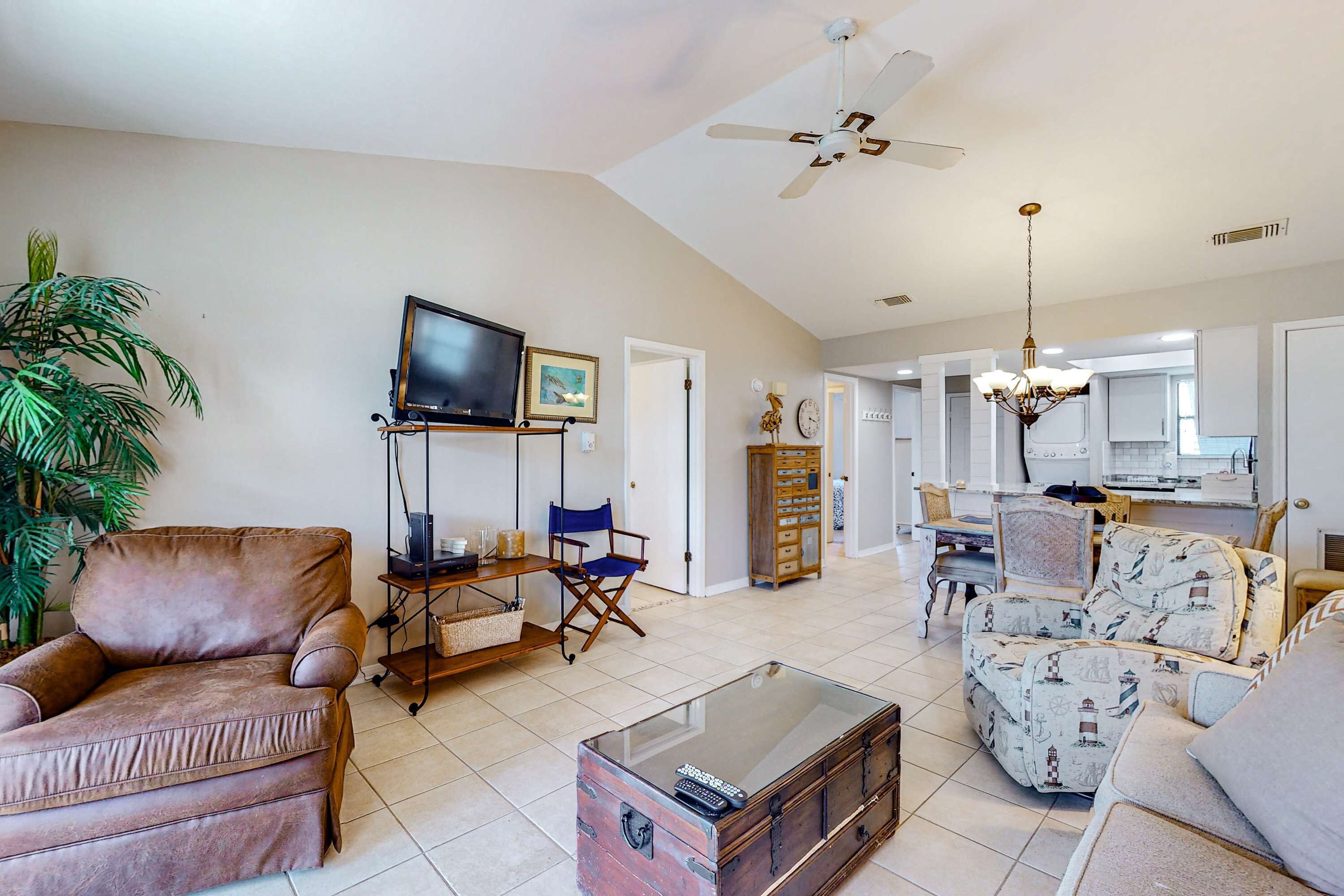 A spacious living room with a brown leather armchair, a glass coffee table, a patterned sofa, a television, and a ceiling fan.