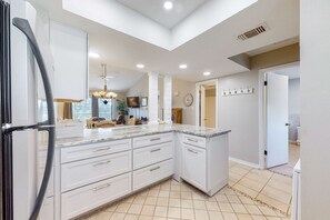 A modern kitchen with white cabinetry, a large island, and a dining area with a chandelier. - Enchanting condo with pool, balcony, central AC, & washer,dryer (Orange Beach)
