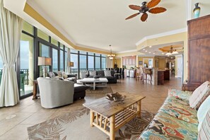 A spacious living area with a high ceiling, large windows, and a view of the outdoors. - Gulf-front corner condo with stunning view, balcony, pools, hot tubs, & gym (Orange Beach)