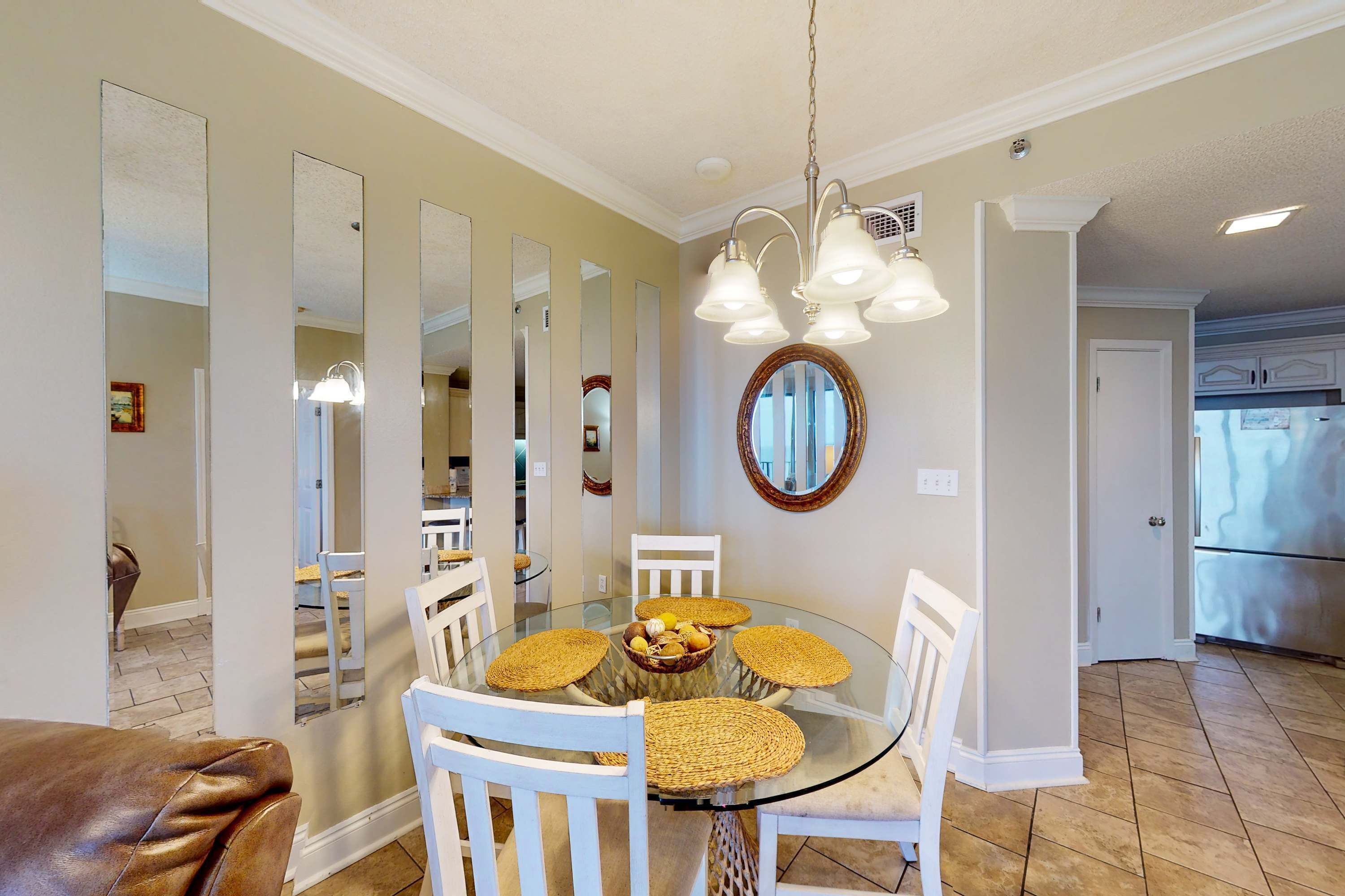 Un coin repas avec une table en verre entourée de chaises blanches, un lustre et un miroir au mur.