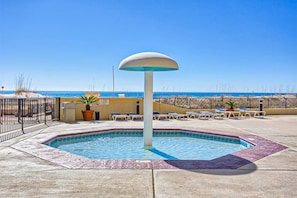 A swimming pool with a central fountain, surrounded by lounge chairs and a fence, with a beach and ocean in the background. - Beachfront condo with Gulf views, balcony, pools, hot tubs & sports courts (Orange Beach)